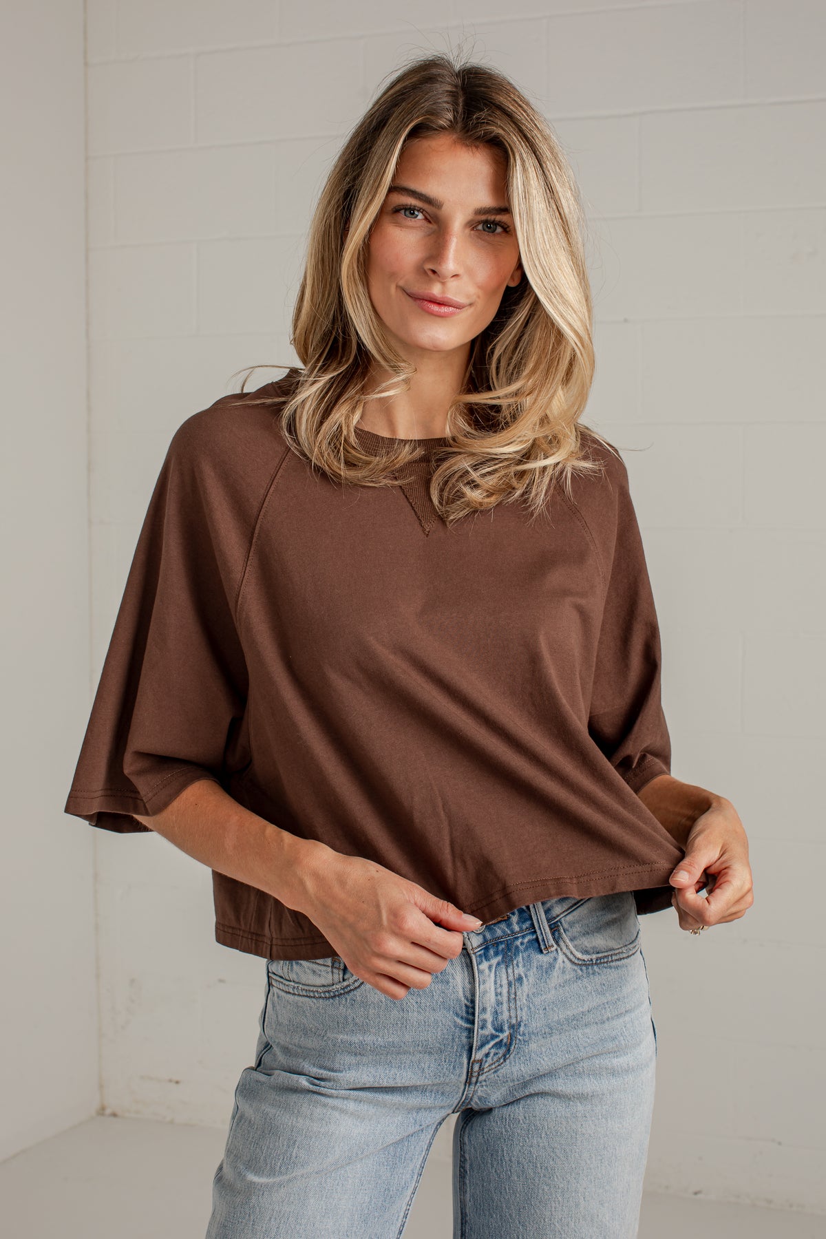 A woman wearing a brown short-sleeve top with wide draped sleeves and light-wash blue jeans, standing against a white brick wall.