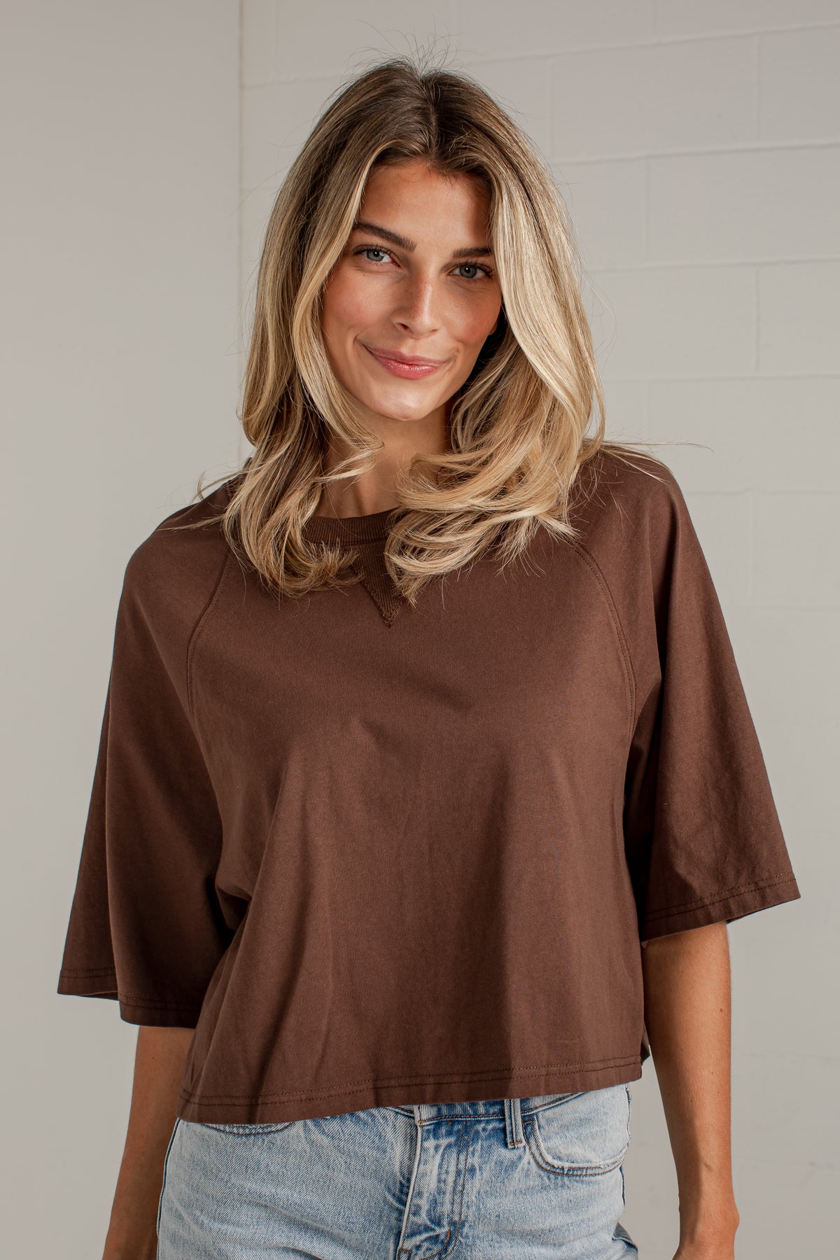 A woman with blonde hair wearing a brown short-sleeve crop top and light blue jeans, standing against a light gray brick wall.