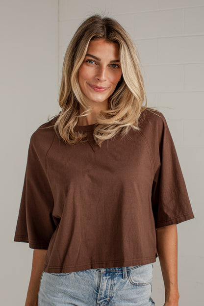 A woman with blonde hair wearing a brown short-sleeve crop top and light blue jeans, standing against a light gray brick wall.