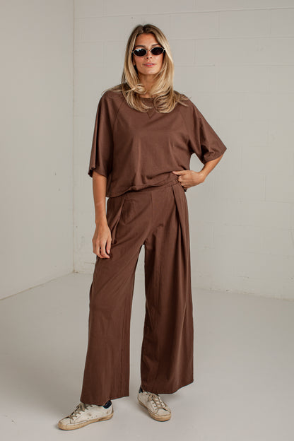 A woman modeling a matching brown two-piece outfit consisting of a loose short-sleeve top and high-waisted wide-leg pants, styled with beige sneakers and sunglasses in a bright studio setting.