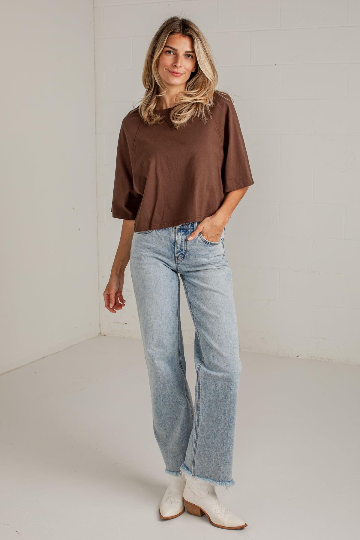 A woman modeling a brown short-sleeve top, light-wash wide-leg jeans, and white ankle boots in a bright, minimalist studio setting.