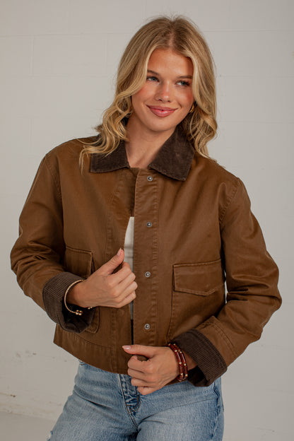A woman wearing a brown jacket with a dark brown corduroy collar and cuffs, paired with light blue jeans, standing against a light gray wall.