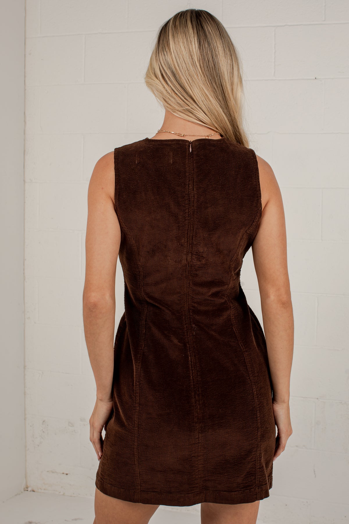 A woman viewed from the back wearing a sleeveless brown mini dress with a textured suede-like finish, standing against a white brick wall.