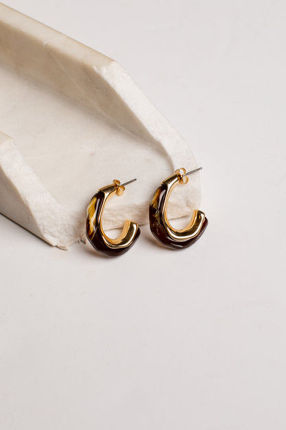 A pair of gold and dark brown tortoiseshell hoop earrings resting on a white textured stone prop against a light gray background.