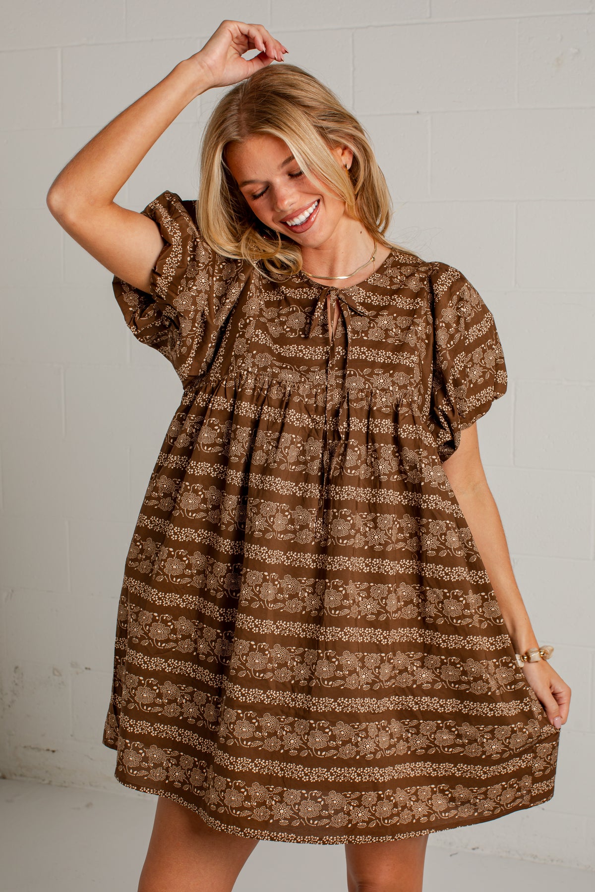 A woman wearing a brown short-sleeve dress with a beige floral horizontal pattern, standing against a white painted brick wall.