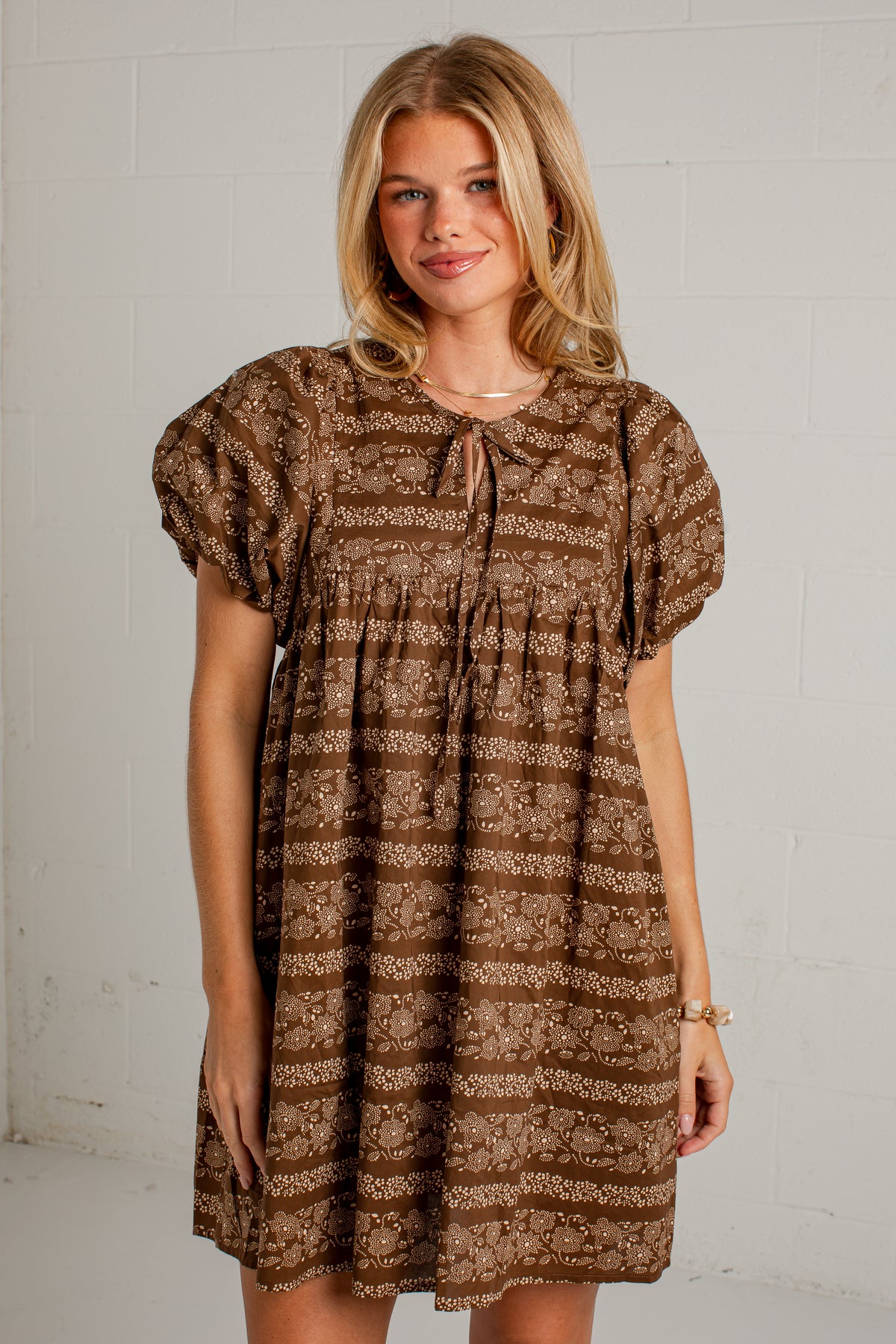 A woman modeling a brown and beige patterned short-sleeve dress with a tie-neck detail, standing against a white brick wall.