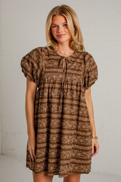 A woman modeling a brown and beige patterned short-sleeve dress with a tie-neck detail, standing against a white brick wall.