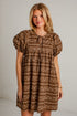 A woman modeling a brown and beige patterned short-sleeve dress with a tie-neck detail, standing against a white brick wall.