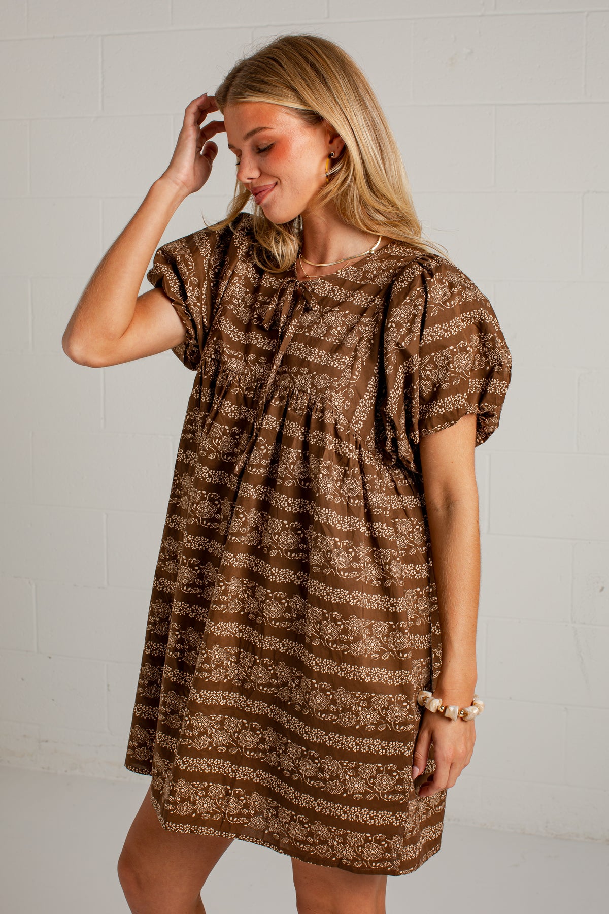 A woman modeling a brown and beige patterned short-sleeve dress against a white brick wall background.