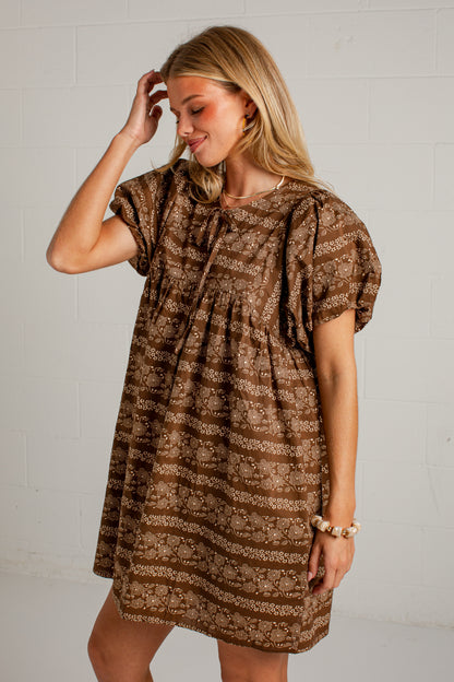 A woman modeling a brown and beige patterned short-sleeve dress against a white brick wall background.