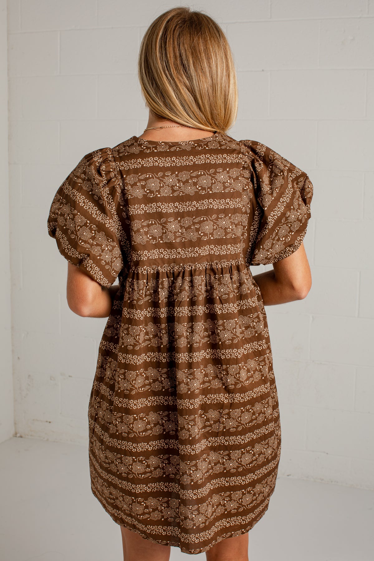 A woman viewed from behind wearing a brown and beige patterned puff-sleeve dress against a white brick wall.