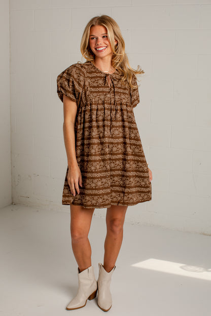 A woman modeling a brown and beige patterned short-sleeve dress with a tie neckline, paired with cream ankle boots, standing in a bright white studio setting.