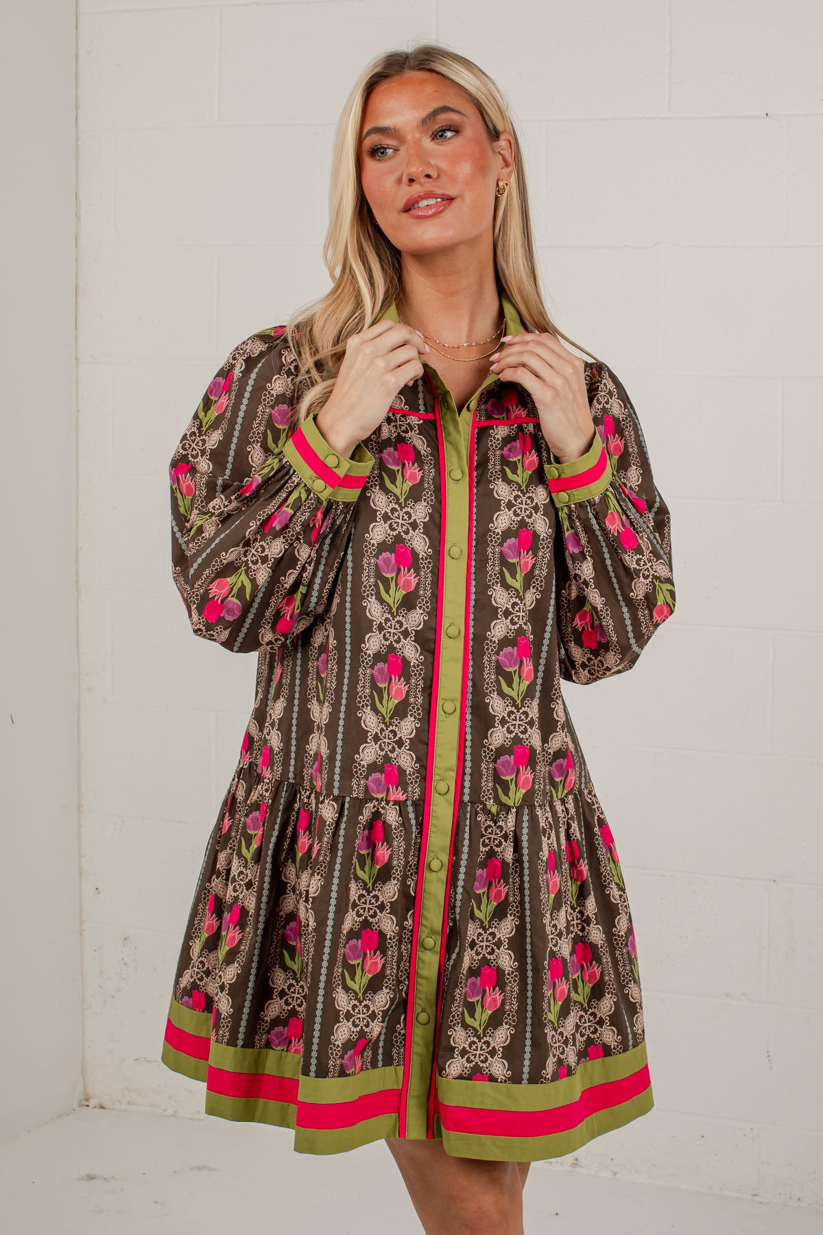 A woman wearing a brown patterned long-sleeve shirt dress with pink and green accents, standing against a white brick wall.