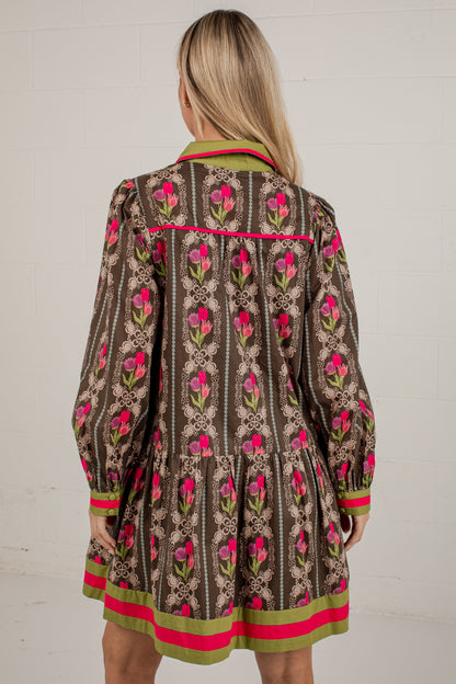 A woman viewed from behind wearing a brown patterned mini dress with pink and green floral motifs and contrasting trim, standing against a white brick wall.