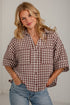 A woman wearing a brown and white gingham check button-down shirt with rolled-up sleeves and blue jeans, standing against a white brick wall.