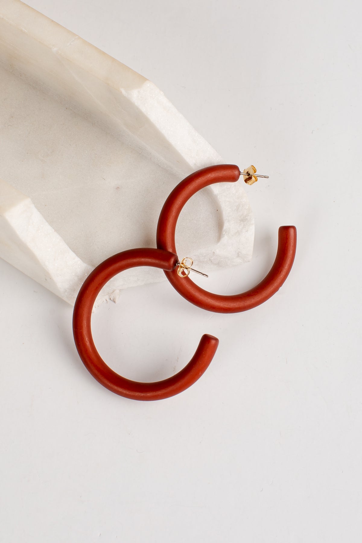 A pair of chunky, matte terracotta-colored hoop earrings with gold post backings, displayed on a white marble prop against a clean white background.