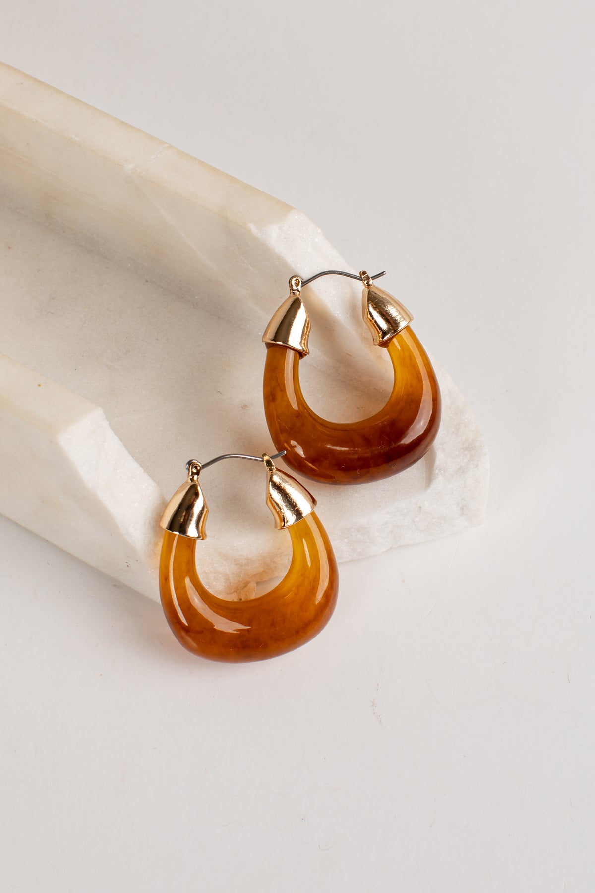 A pair of crescent-shaped hoop earrings made of glossy amber-brown translucent material with gold-toned metal tops and posts, displayed on a white stone slab against a light neutral background.