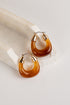 A pair of crescent-shaped hoop earrings made of glossy amber-brown translucent material with gold-toned metal tops and posts, displayed on a white stone slab against a light neutral background.