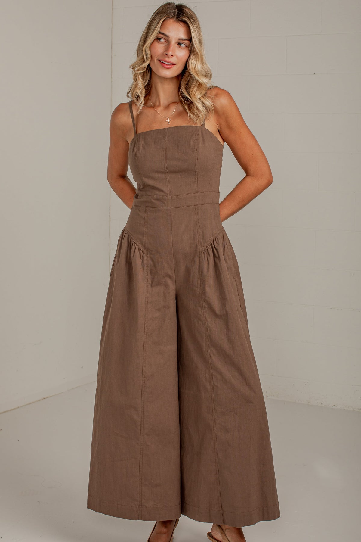 A woman modeling a brown wide-leg strapless jumpsuit in a bright, minimalist studio setting.