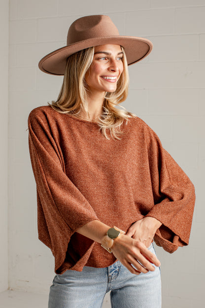 A woman wearing a rust-brown knit sweater, a wide-brimmed brown hat, and light-wash blue jeans, standing against a white brick wall.