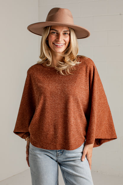 A woman wearing a rust-brown knit sweater, a wide-brimmed brown hat, and light-wash blue jeans, standing against a white brick wall.