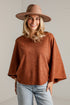 A woman wearing a rust-brown knit sweater, a wide-brimmed brown hat, and light-wash blue jeans, standing against a white brick wall.