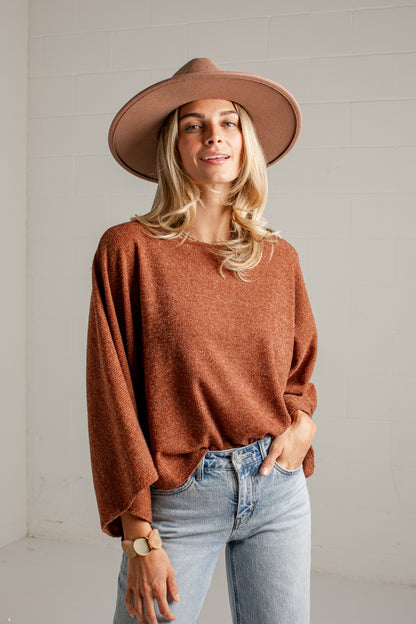 A woman wearing a rust-brown knit sweater, a wide-brimmed brown hat, and light-wash blue jeans, standing against a white brick wall.