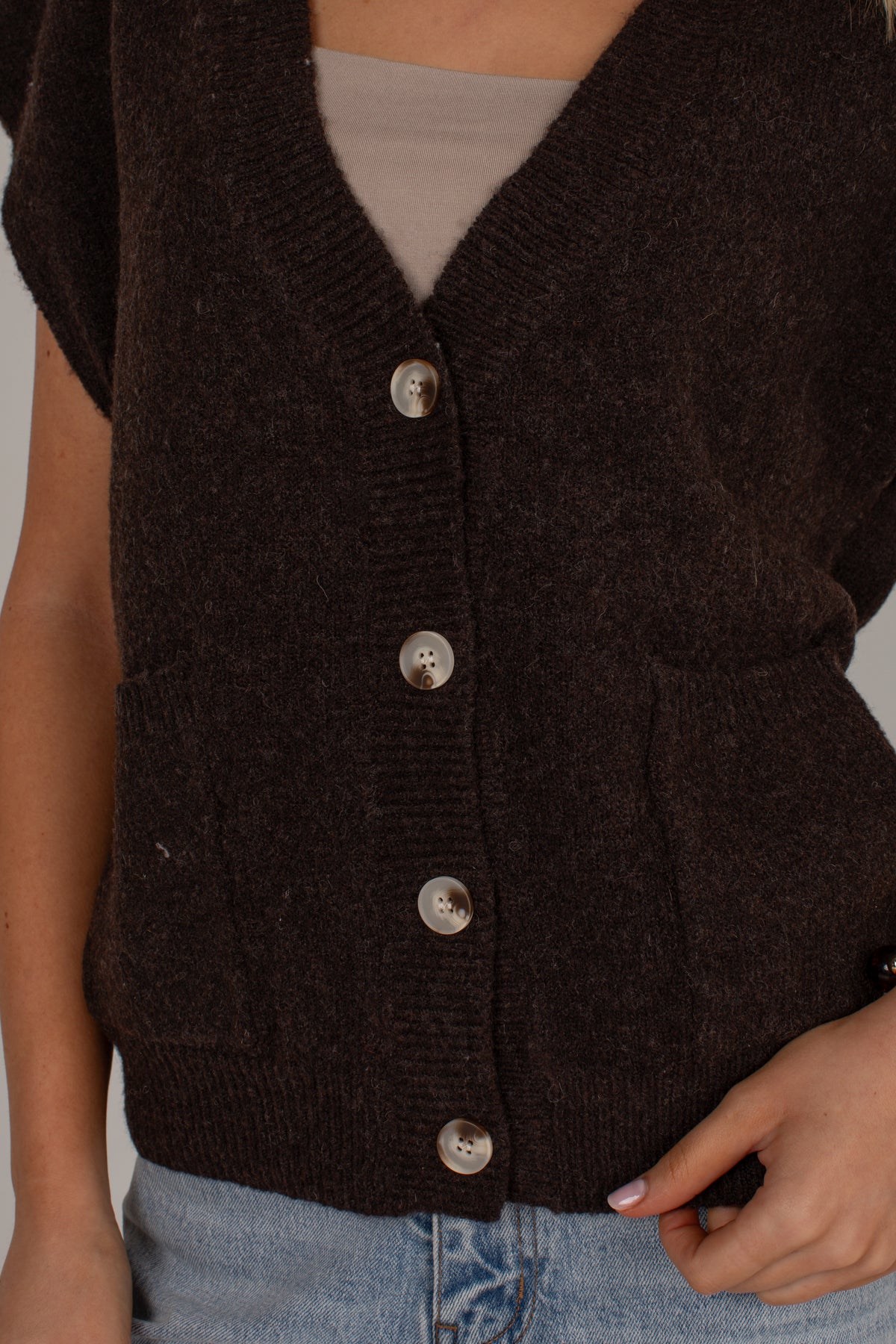 A close-up of a person wearing a dark brown knit cardigan vest over a beige top and light blue jeans. The cardigan features a V-neck, silver buttons, and side pockets.