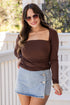 Woman wearing a brown long-sleeve top and denim skirt on a porch.