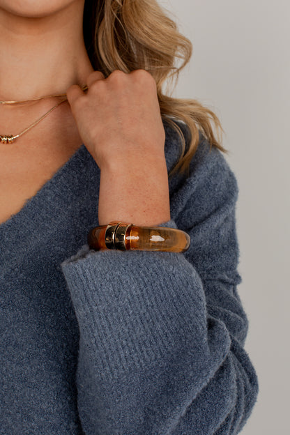A close-up of a woman wearing a chunky amber-brown translucent bangle bracelet with silver-toned metal accents on her wrist, paired with a blue knit sweater.