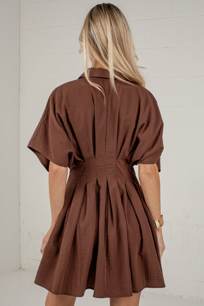 A woman viewed from behind wearing a brown short-sleeve mini dress with a gathered waist and pleated back detail, standing against a white paneled wall.