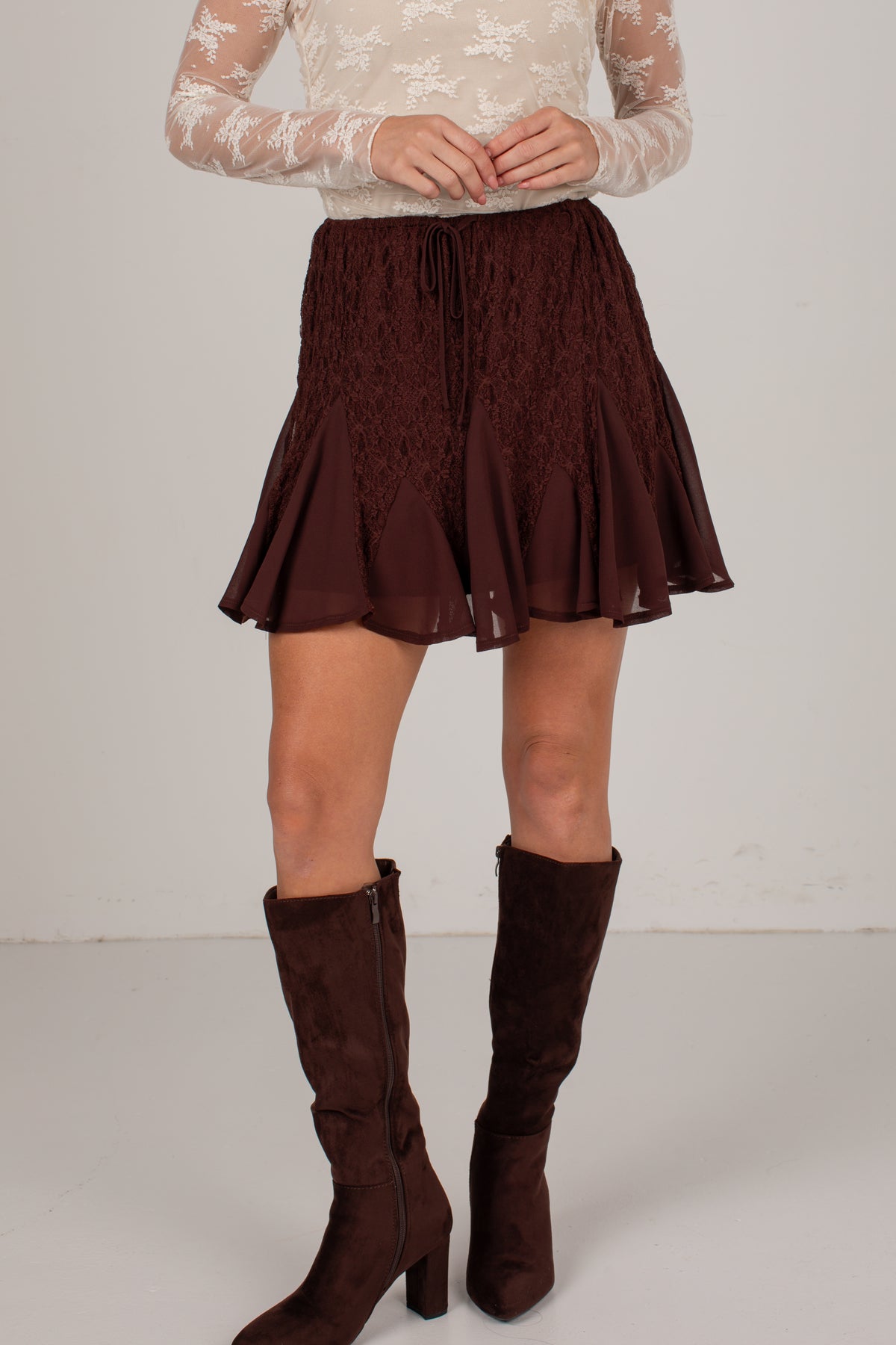 A model wearing a dark brown lace and mesh mini skirt with a drawstring waist, paired with matching dark brown suede knee-high boots and a cream lace long-sleeve top, set against a plain light gray studio background.