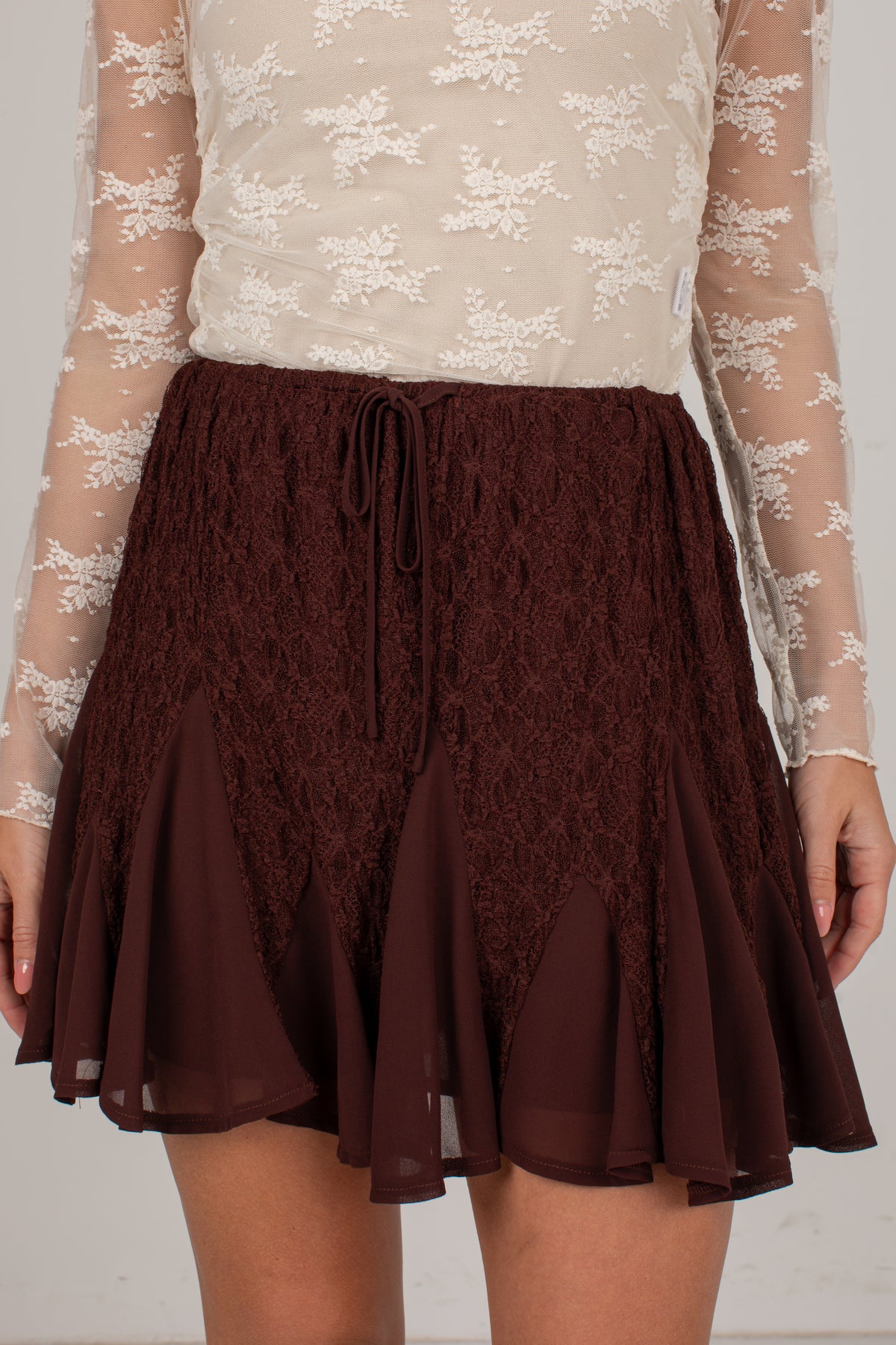 A close-up of a model wearing a dark brown lace and chiffon flared skirt with a drawstring waist, paired with a cream lace long-sleeve top.