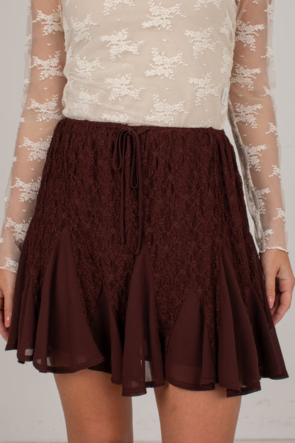 A close-up of a model wearing a dark brown lace and chiffon flared skirt with a drawstring waist, paired with a cream lace long-sleeve top.