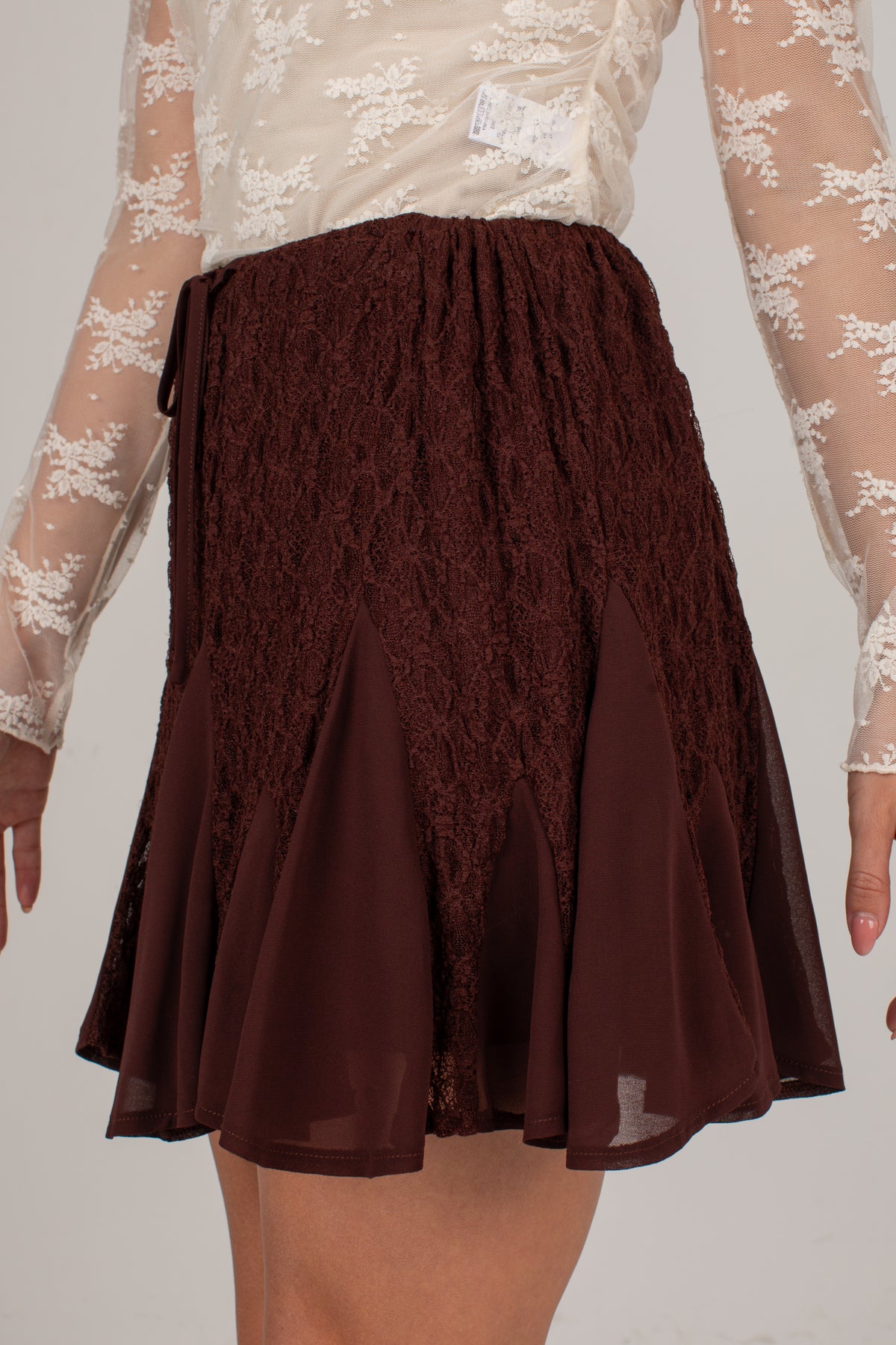 A dark brown skirt featuring a textured lace front panel and a flowing chiffon flared hem, shown on a model wearing a sheer cream lace top.