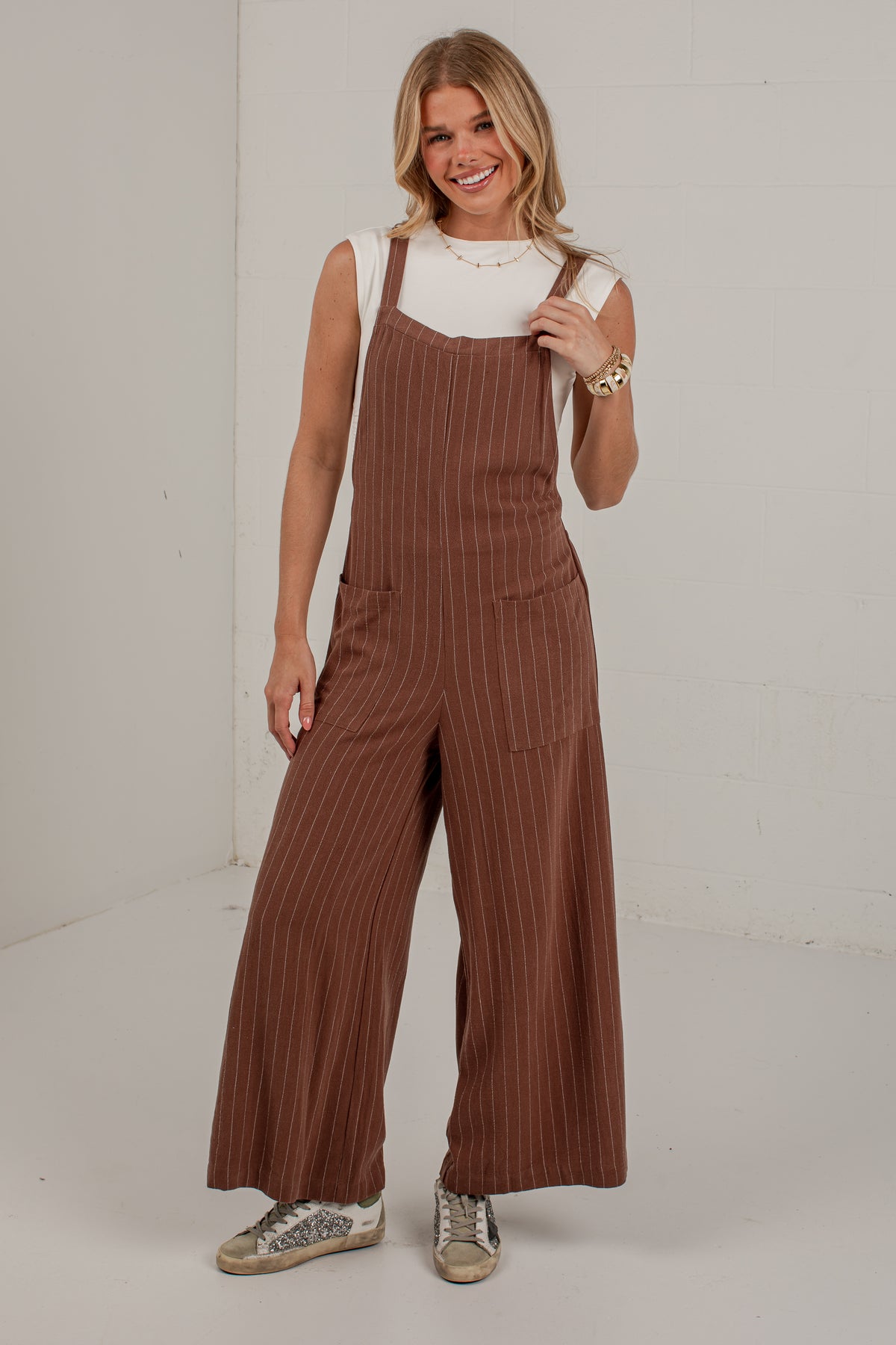 A woman modeling a pair of brown pinstriped wide-leg overall jumpsuits over a white sleeveless top, standing in a white studio setting.