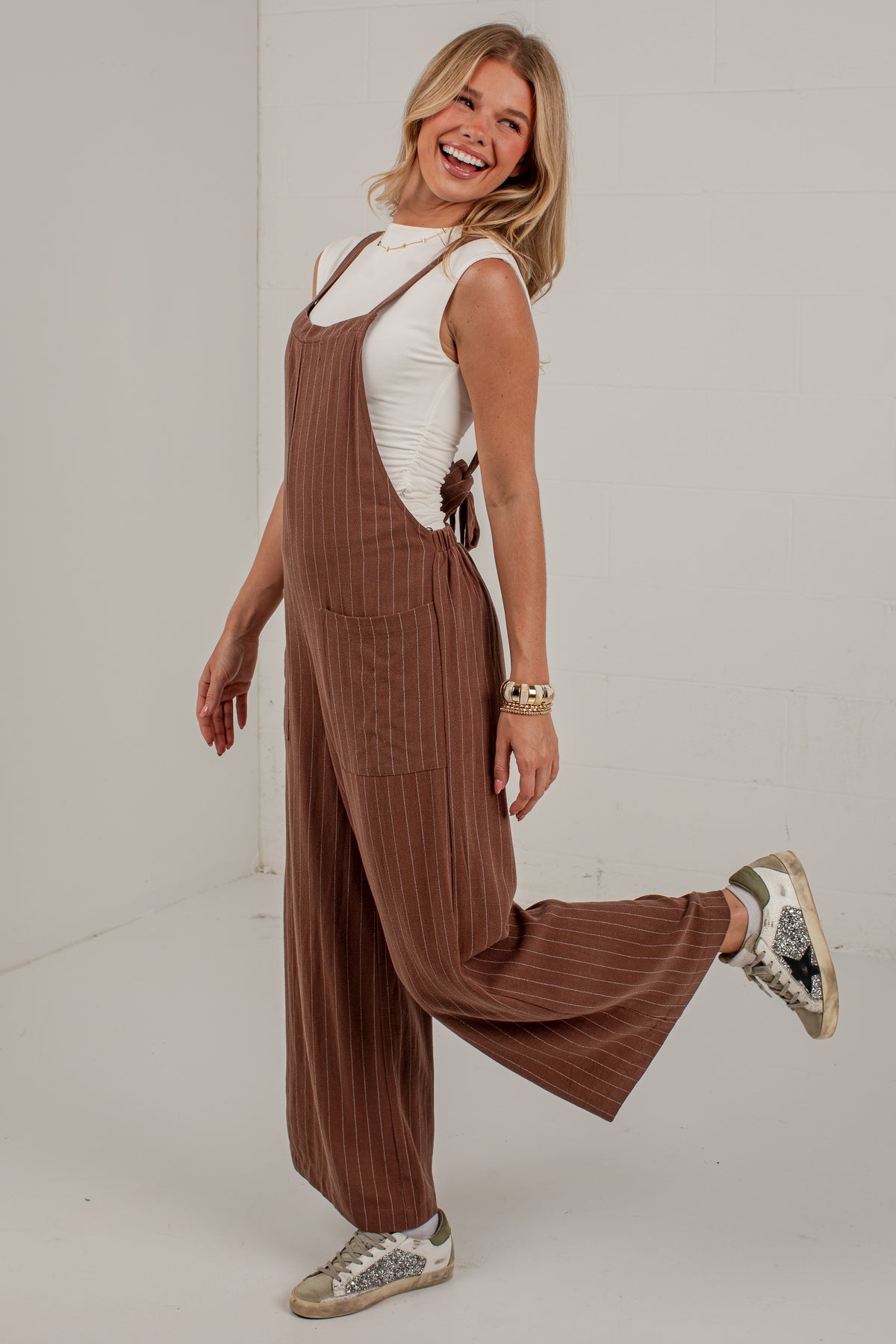 A woman modeling a brown pinstriped corduroy jumpsuit with wide legs, styled with a white sleeveless top and distressed sneakers in a bright studio setting.