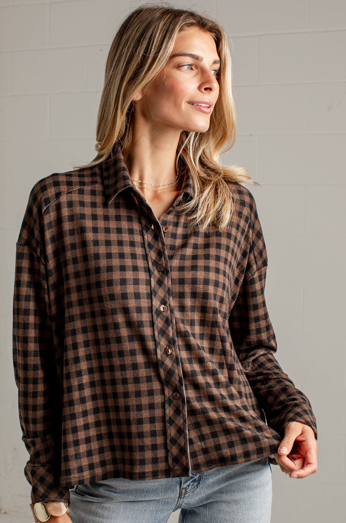 A woman wearing a brown and black checkered long-sleeve button-up shirt with light blue jeans, standing against a light gray brick wall.