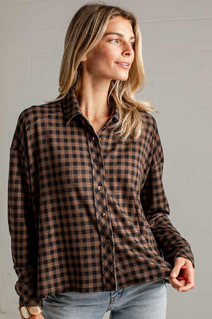 A woman wearing a brown and black checkered long-sleeve button-up shirt with light blue jeans, standing against a light gray brick wall.