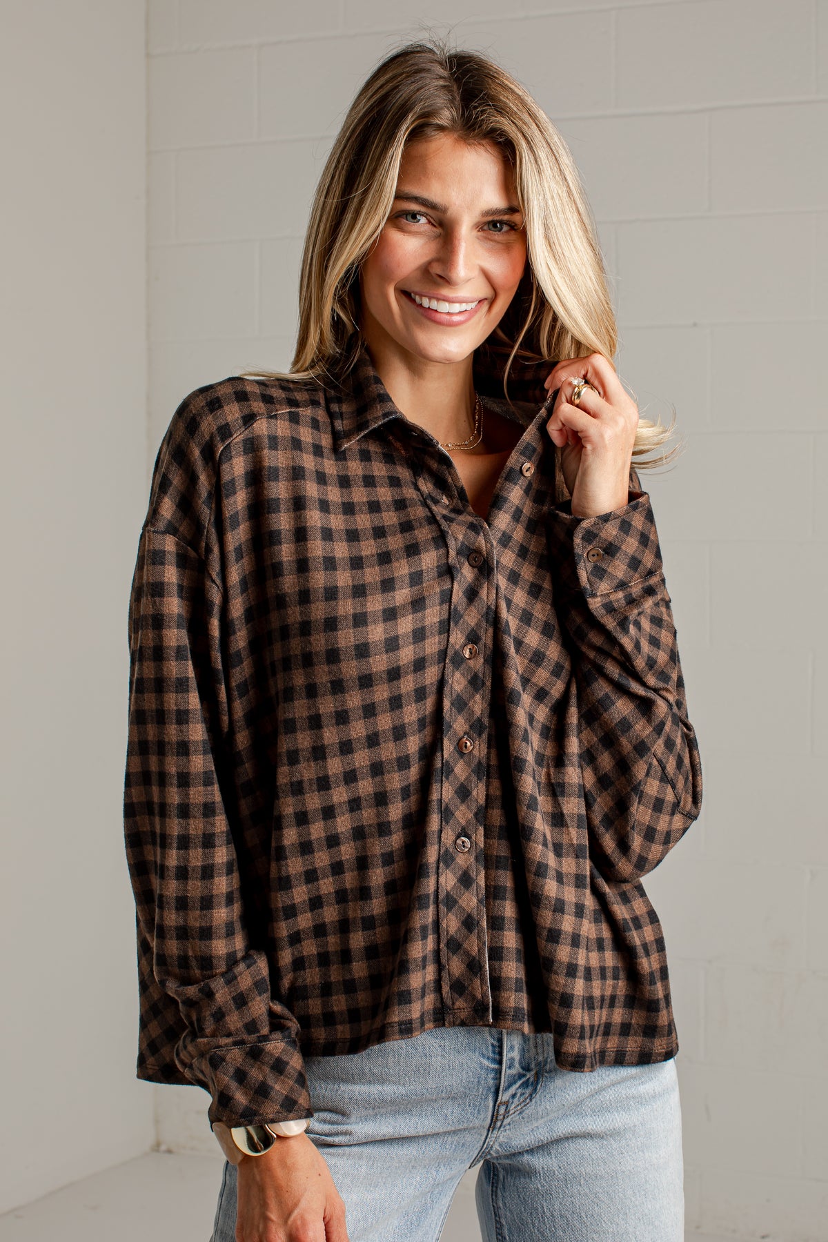 A woman wearing a brown and black checkered plaid button-down shirt with light blue jeans, standing against a white brick wall.