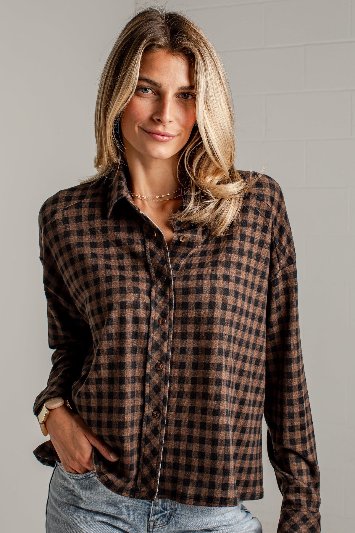 A woman wearing a brown and black checkered long-sleeve button-up shirt with light blue jeans, standing against a light gray brick wall.