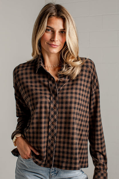 A woman wearing a brown and black checkered long-sleeve button-up shirt with light blue jeans, standing against a light gray brick wall.