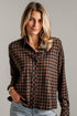 A woman wearing a brown and black checkered long-sleeve button-up shirt with light blue jeans, standing against a light gray brick wall.