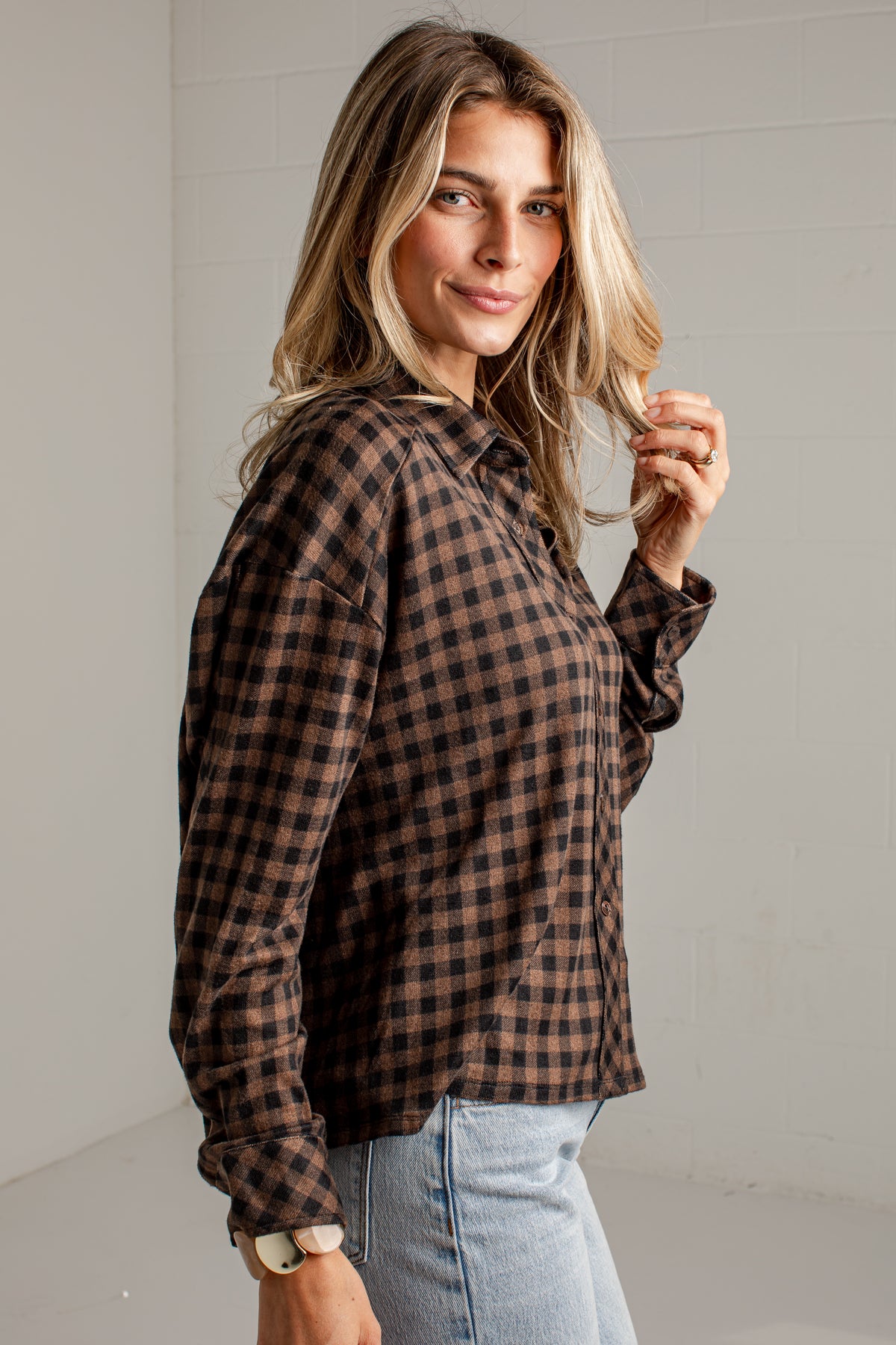 A woman wearing a brown and black checkered flannel shirt with light blue jeans, standing against a white paneled wall.
