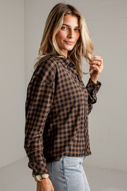 A woman wearing a brown and black checkered flannel shirt with light blue jeans, standing against a white paneled wall.