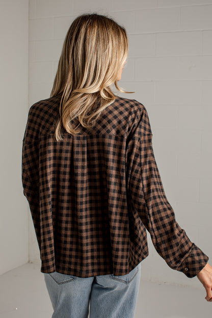 A woman viewed from behind wearing a brown and black buffalo plaid flannel shirt and light blue jeans, standing against a white brick wall.