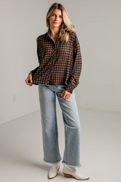 A woman modeling a brown and black checkered long-sleeve button-up shirt, paired with light blue wide-leg jeans and white cowboy boots, standing against a white studio wall.