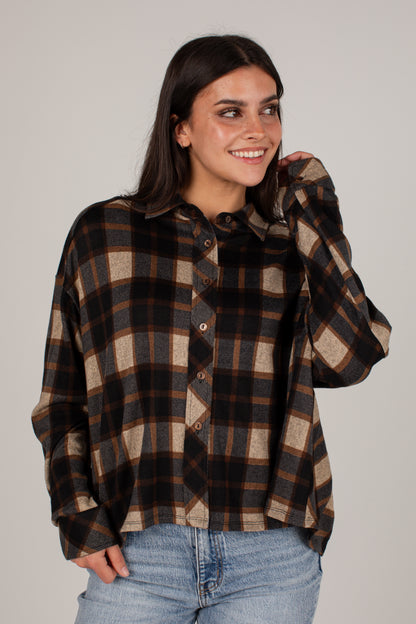 A woman wearing a long-sleeve button-up plaid shirt in brown, black, and beige, paired with light blue jeans, against a solid light gray background.