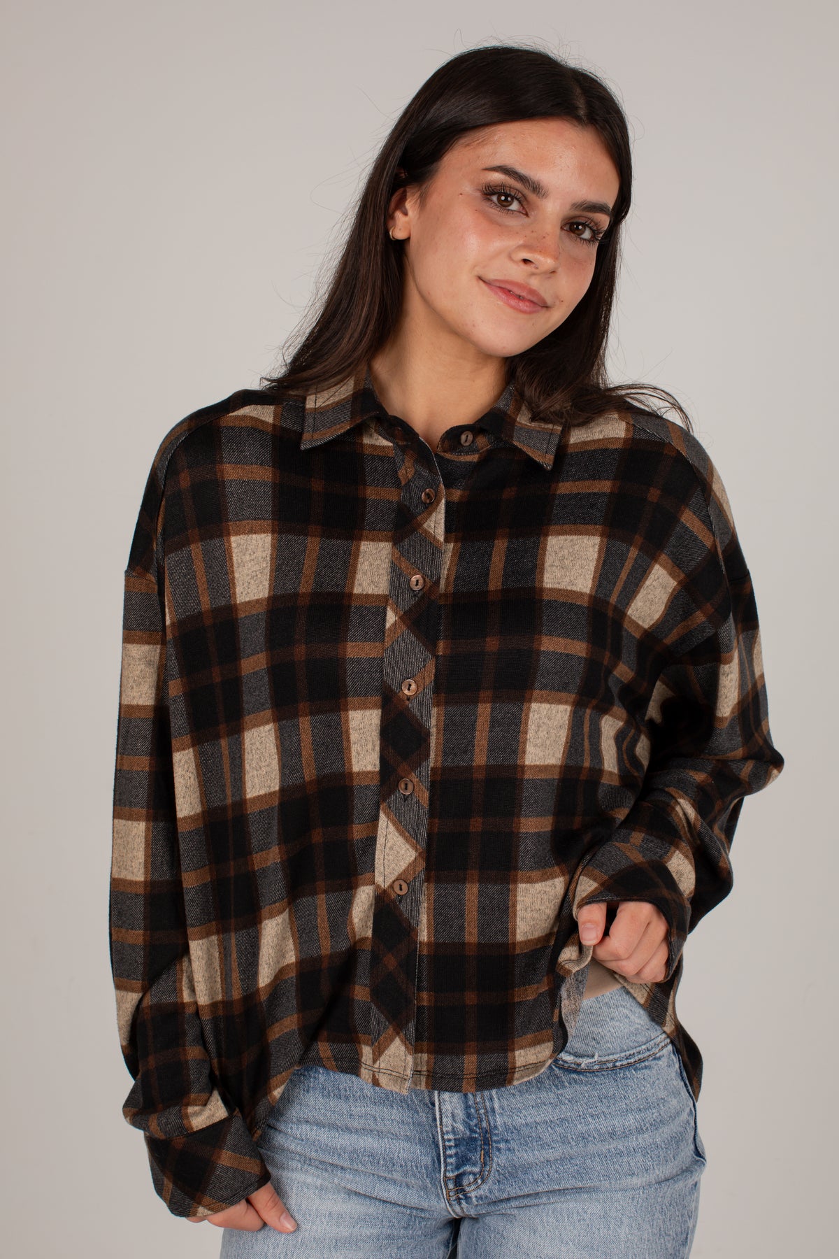 A woman wearing an oversized long-sleeve plaid button-up shirt in brown, black, and beige, paired with light blue jeans, against a plain light gray studio background.
