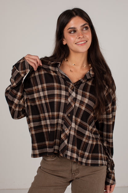A woman modeling a long-sleeve button-up plaid shirt in brown, beige, and black, paired with brown pants, against a plain light gray studio background.